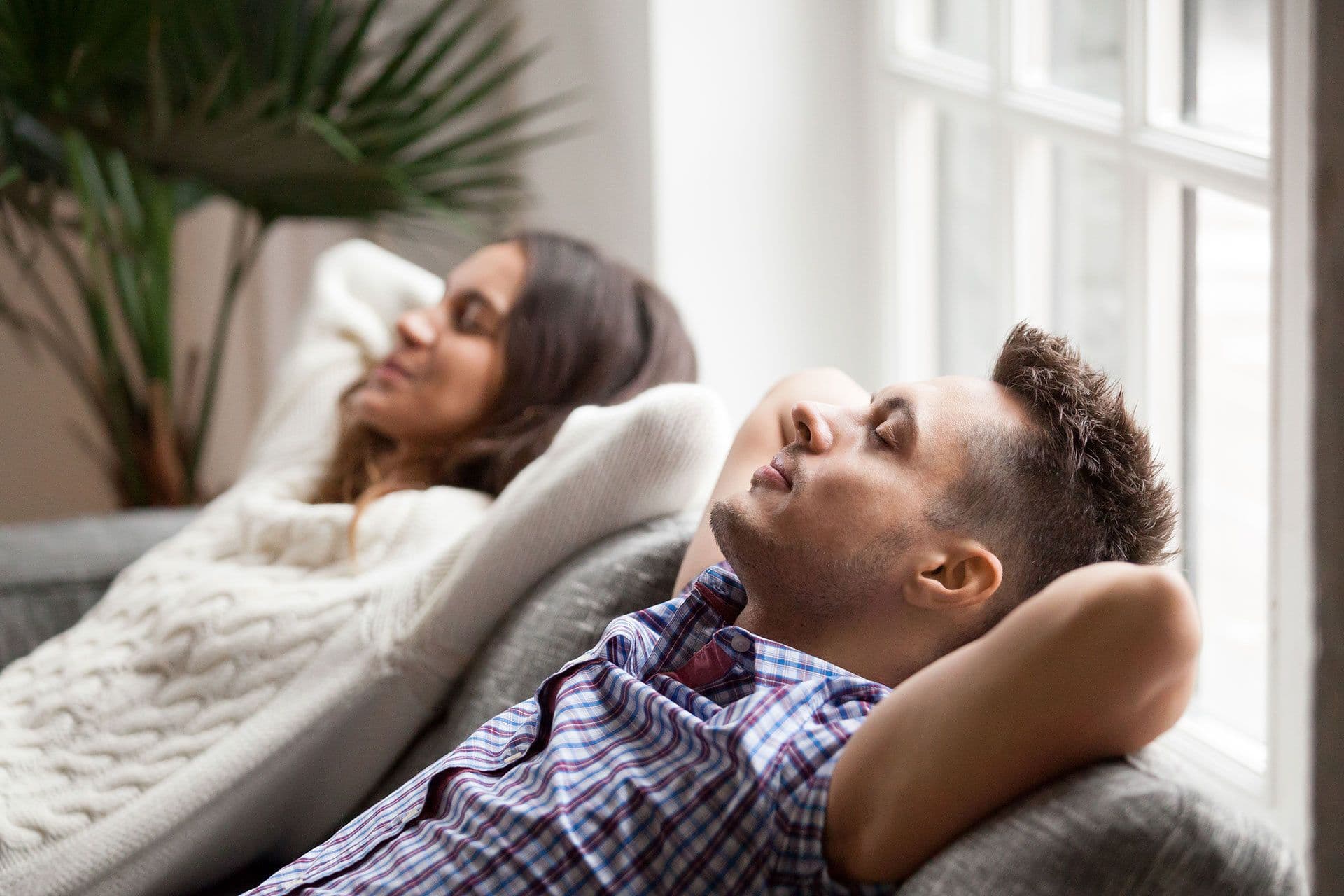 A couple relaxes on a couch with their eyes closed and arms crossed behind their heads.