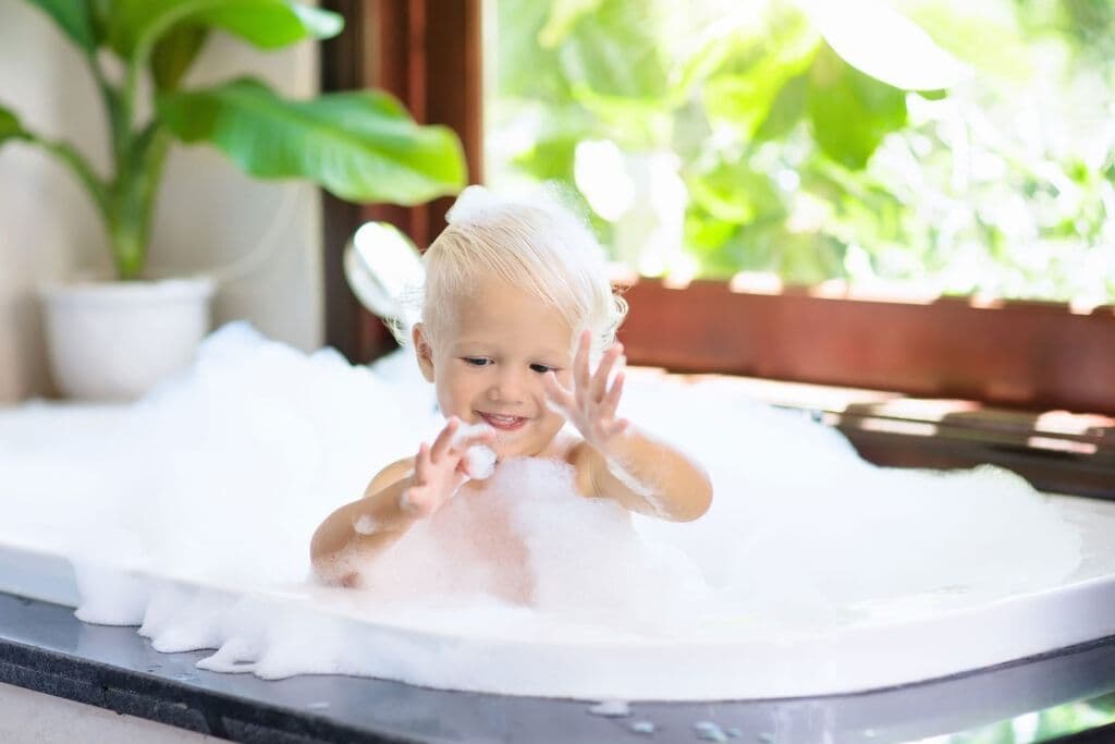 A smiling toddler plays with bubbles in a sunlit bathtub near a window. Lush green plants frame the scene, creating a joyful and serene atmosphere.