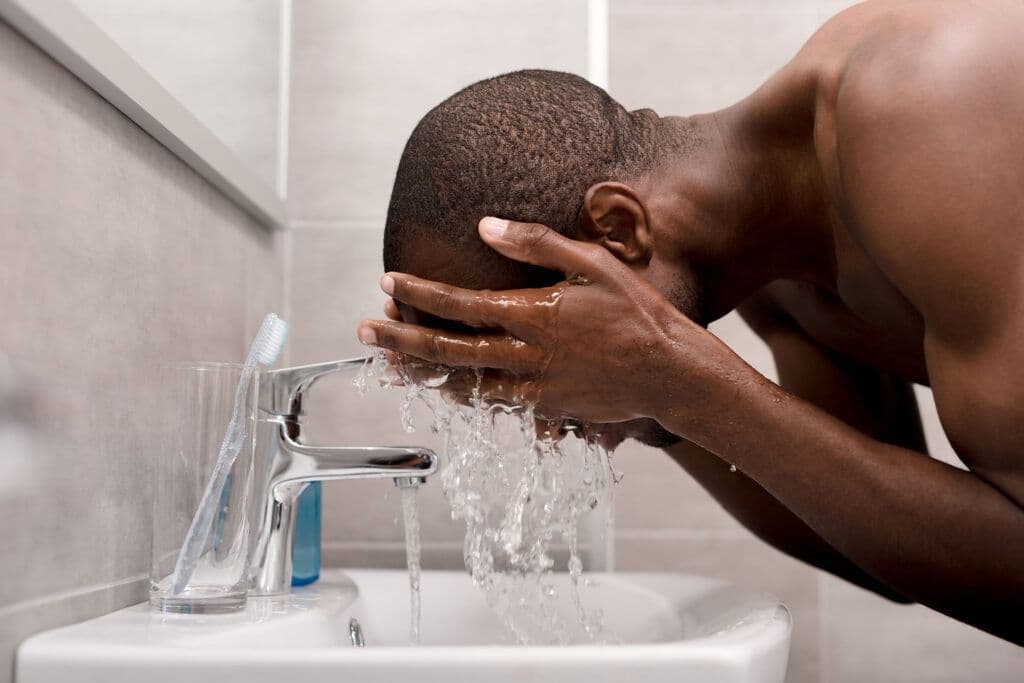 A man leans over a bathroom sink, splashing water onto his face with cupped hands. A toothbrush is visible on the counter beside a modern faucet.